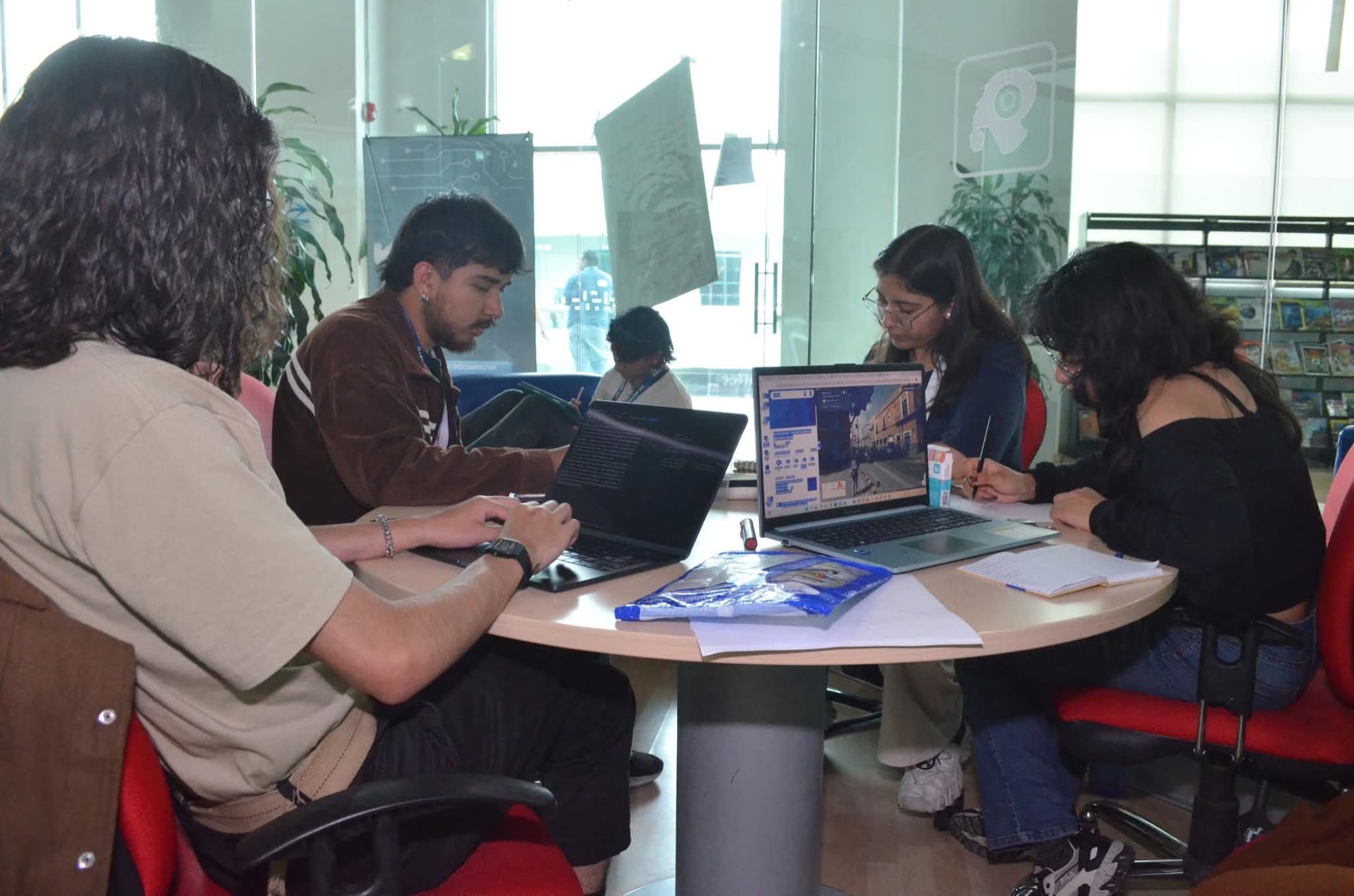 Four students work at a round table with laptops, notes and a bag of materials.