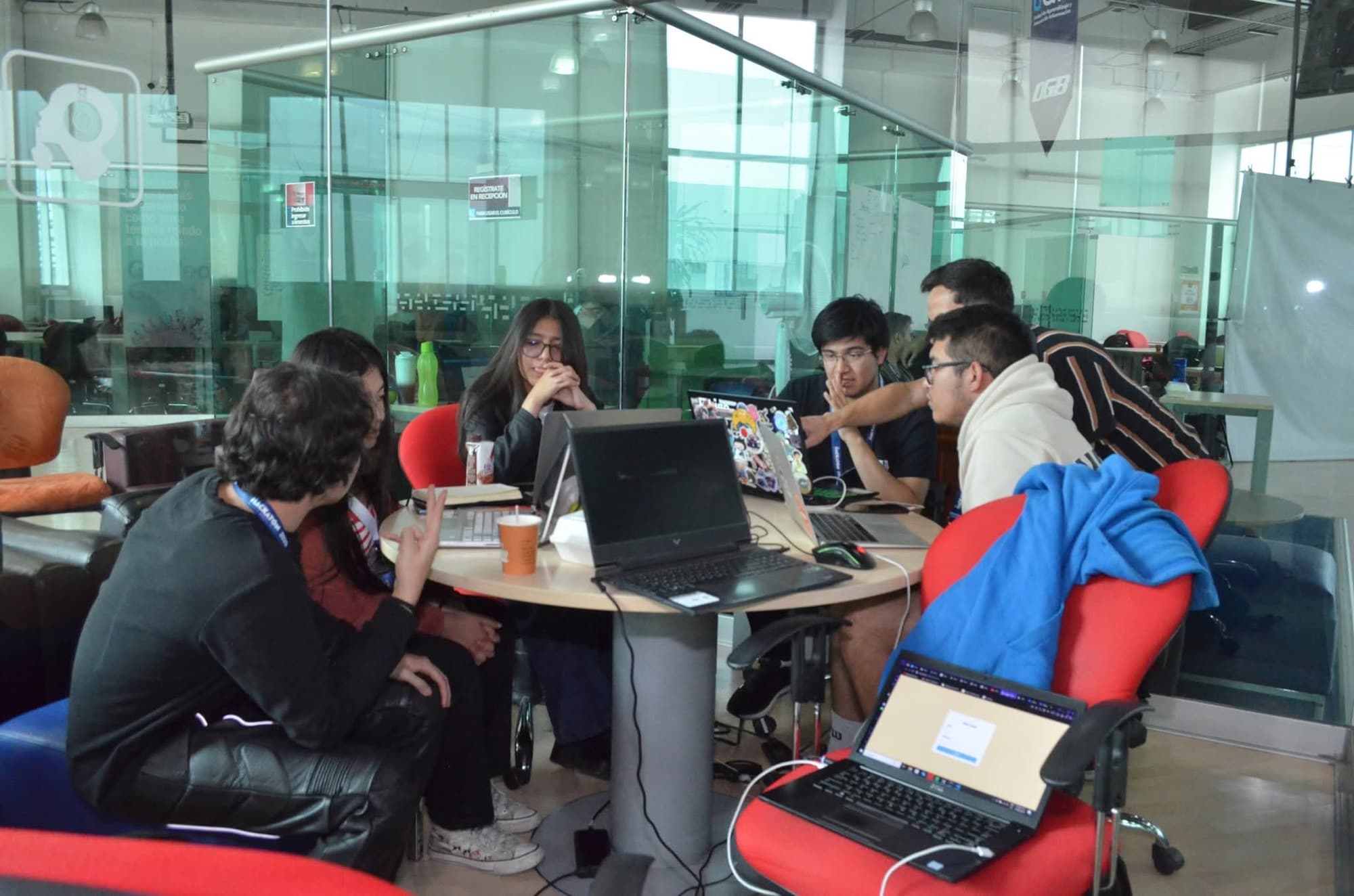 A team of five students works with laptops at a round table inside a glass room.