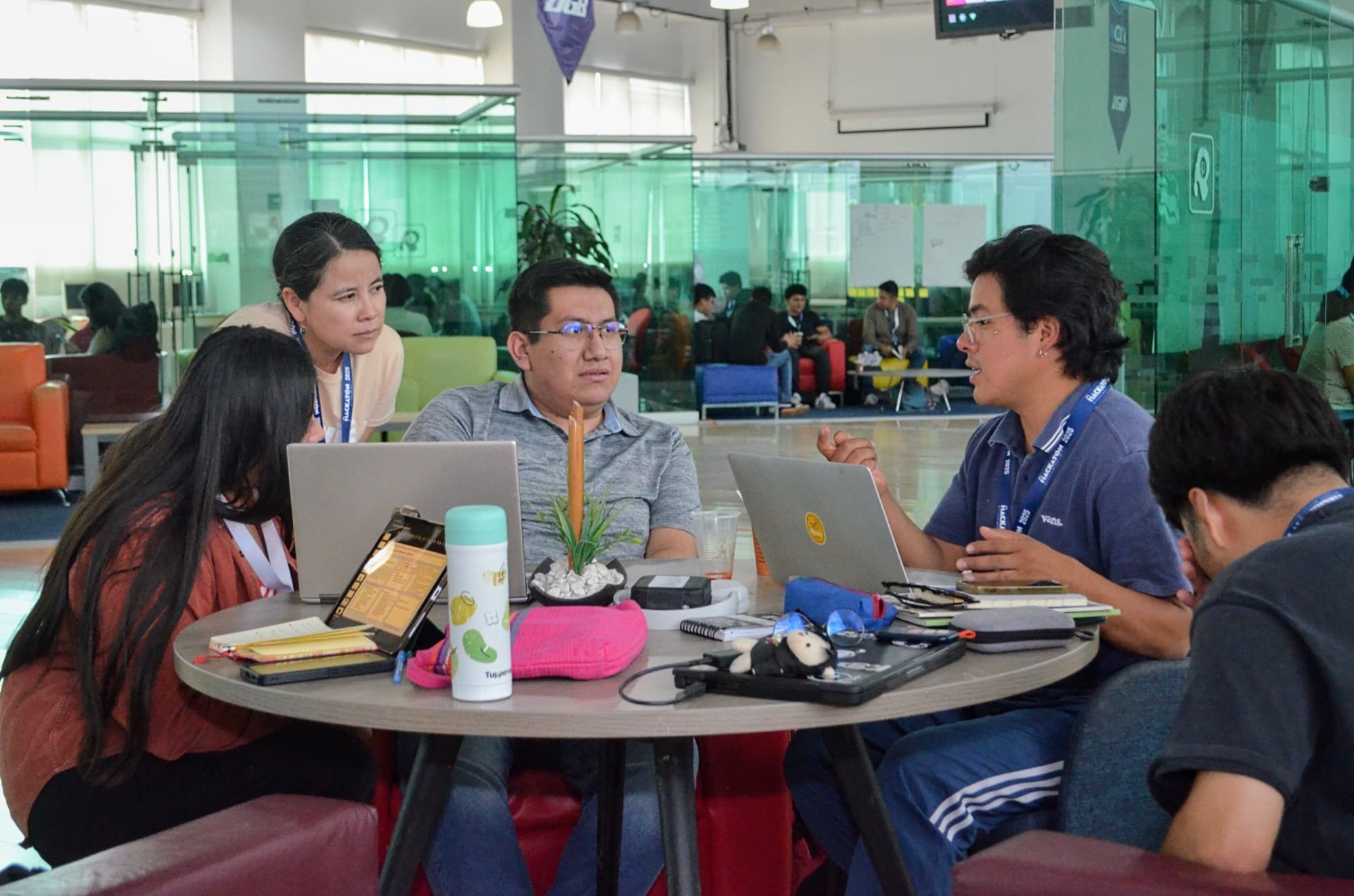 Four students talk around a table with laptops in a glass-walled study room.