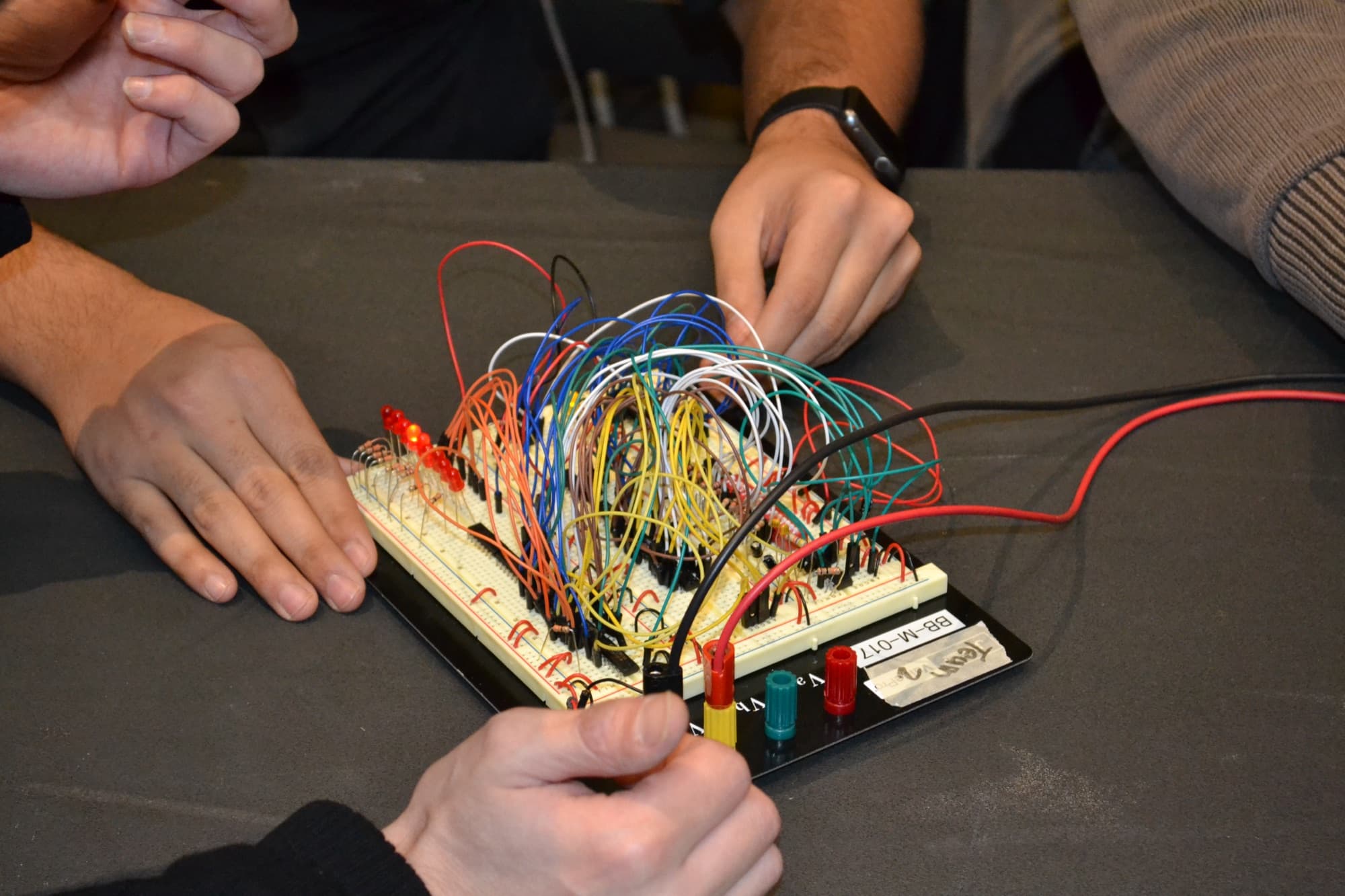 Three students' hands wiring a breadboard packed with colored jumper wires and red LEDs, at a dark table.